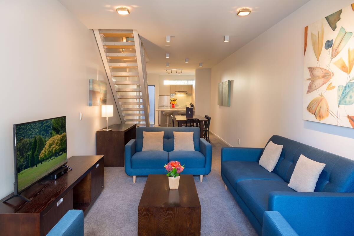 Un salon moderne avec un canapé bleu, une table basse ornée de fleurs, un téléviseur posé sur un meuble en bois et un escalier menant à l’étage supérieur.