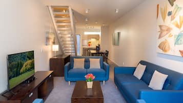 Una sala de estar moderna con un sofá azul, una mesa de centro con flores, un televisor sobre un mueble de madera y una escalera que conduce a un piso superior.
