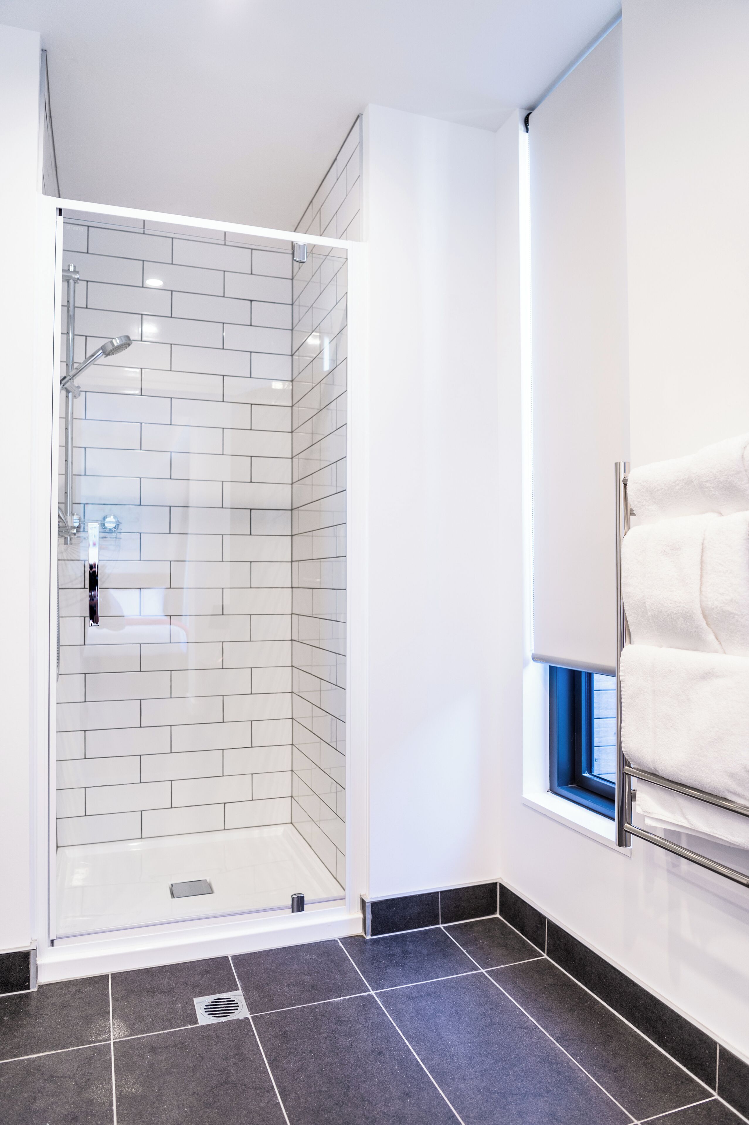 A modern bathroom with a glass-enclosed shower, white tiled walls, a black tiled floor, and a window with a towel rack.