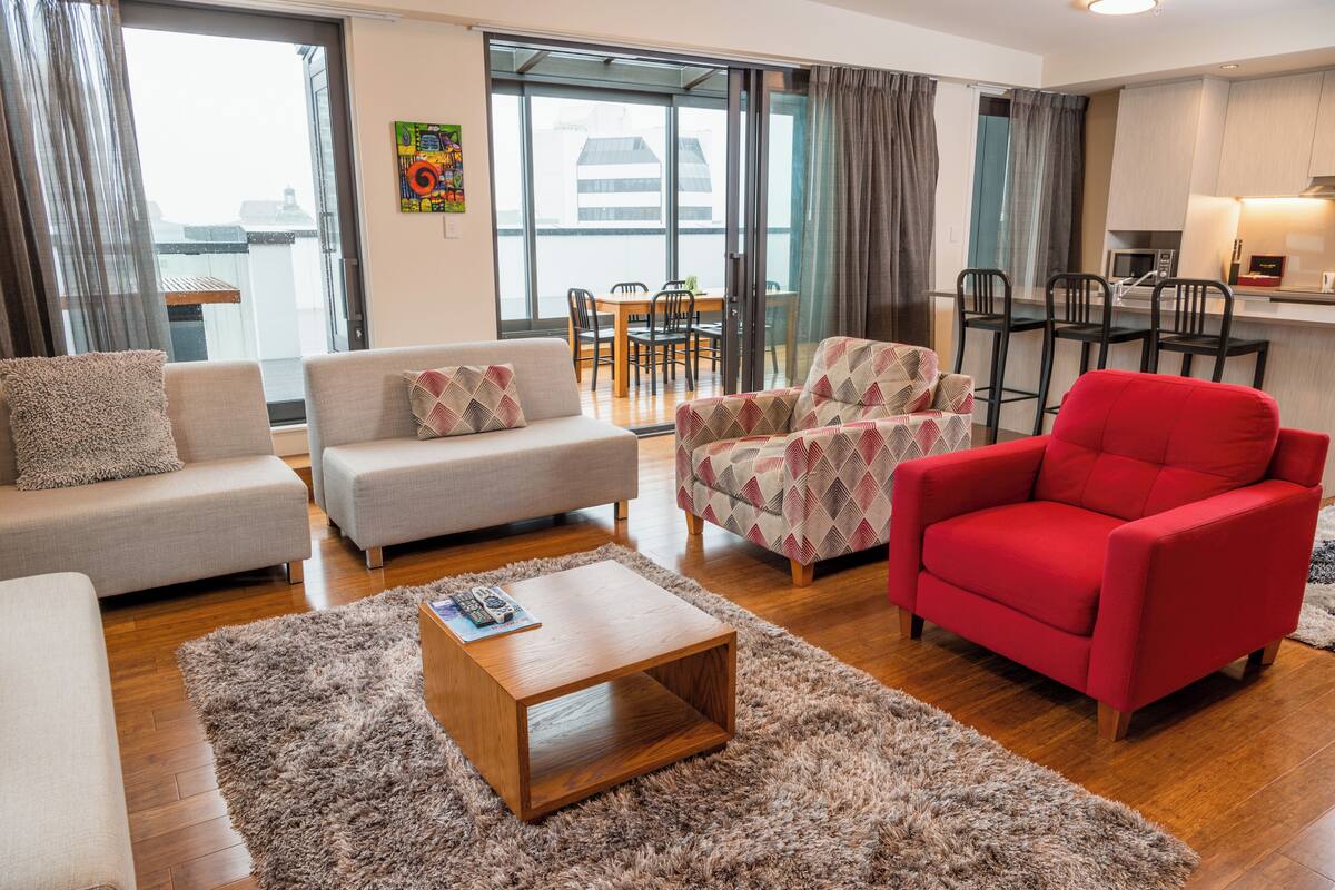 Un salon moderne avec un canapé, un fauteuil et un fauteuil rouge. On y trouve une table à manger avec des chaises, un coin cuisine et une vue à travers la porte vitrée.