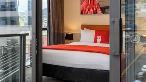 A hotel room with a bed, a window with curtains, a painting on the wall, and a balcony with a glass door. - Boulcott Suites (Wellington)