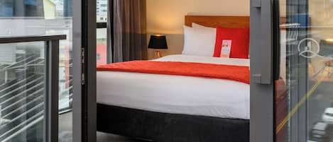 A hotel room with a bed, a window with curtains, a painting on the wall, and a balcony with a glass door.