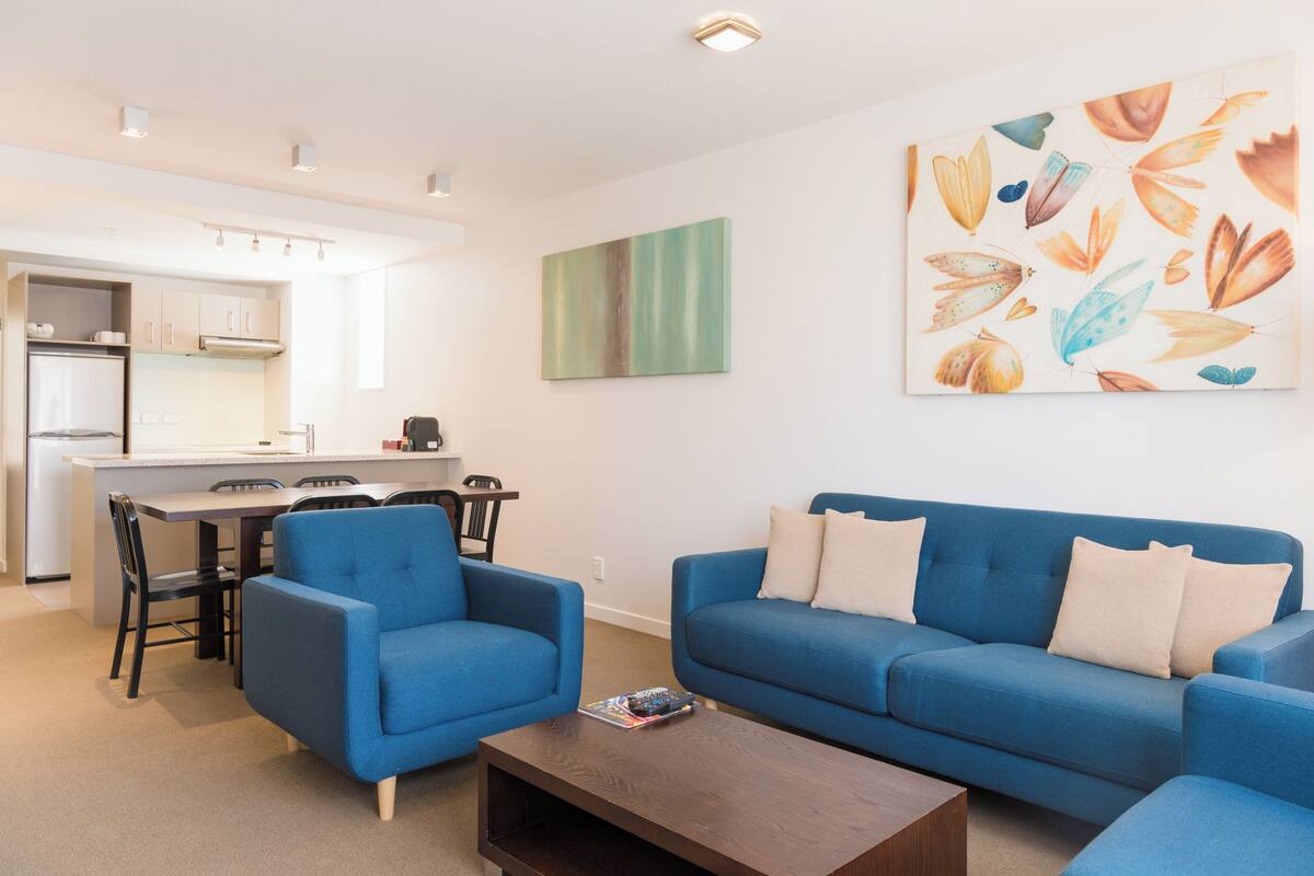 Un salon moderne avec un canapé bleu, des fauteuils assortis et une table basse en bois. On aperçoit à l’arrière-plan un coin cuisine avec des meubles blancs et une table à manger.