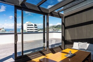 A modern balcony with a wooden table and seating, large glass doors, and a view of buildings and hills.