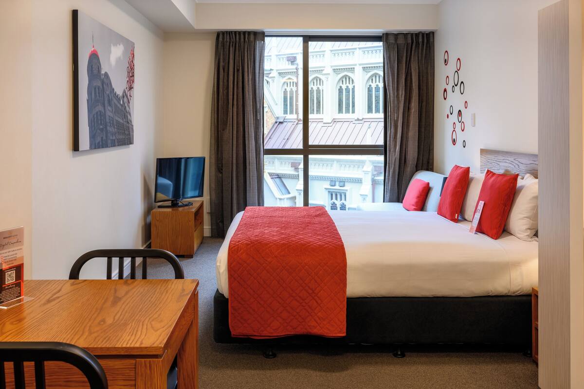 Une chambre d’hôtel avec un grand lit, un bureau, une chaise, une télévision et une fenêtre donnant sur un immeuble.