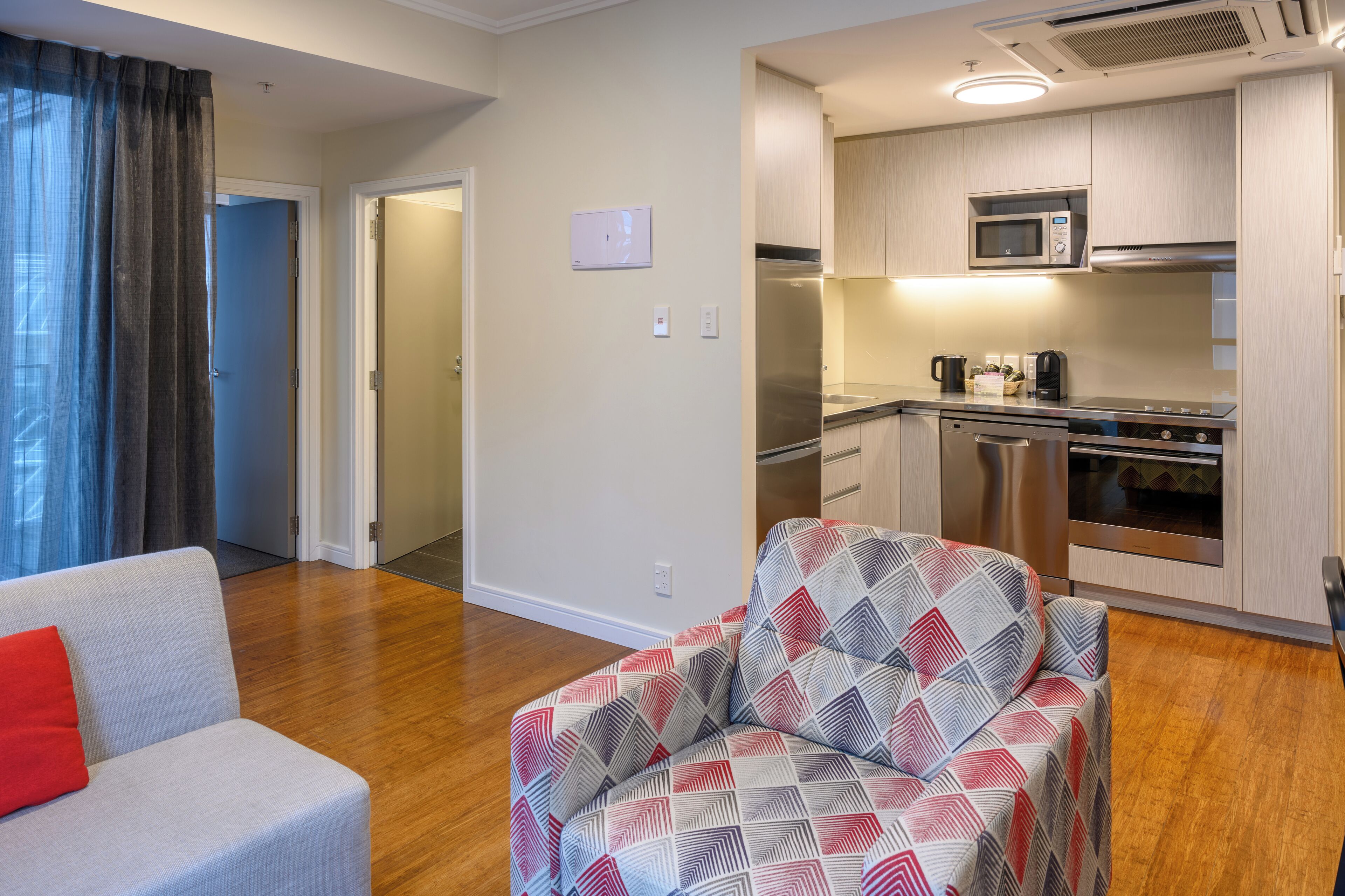 A modern apartment with a living room, a patterned armchair, a kitchen with stainless steel appliances, and a view through a door.