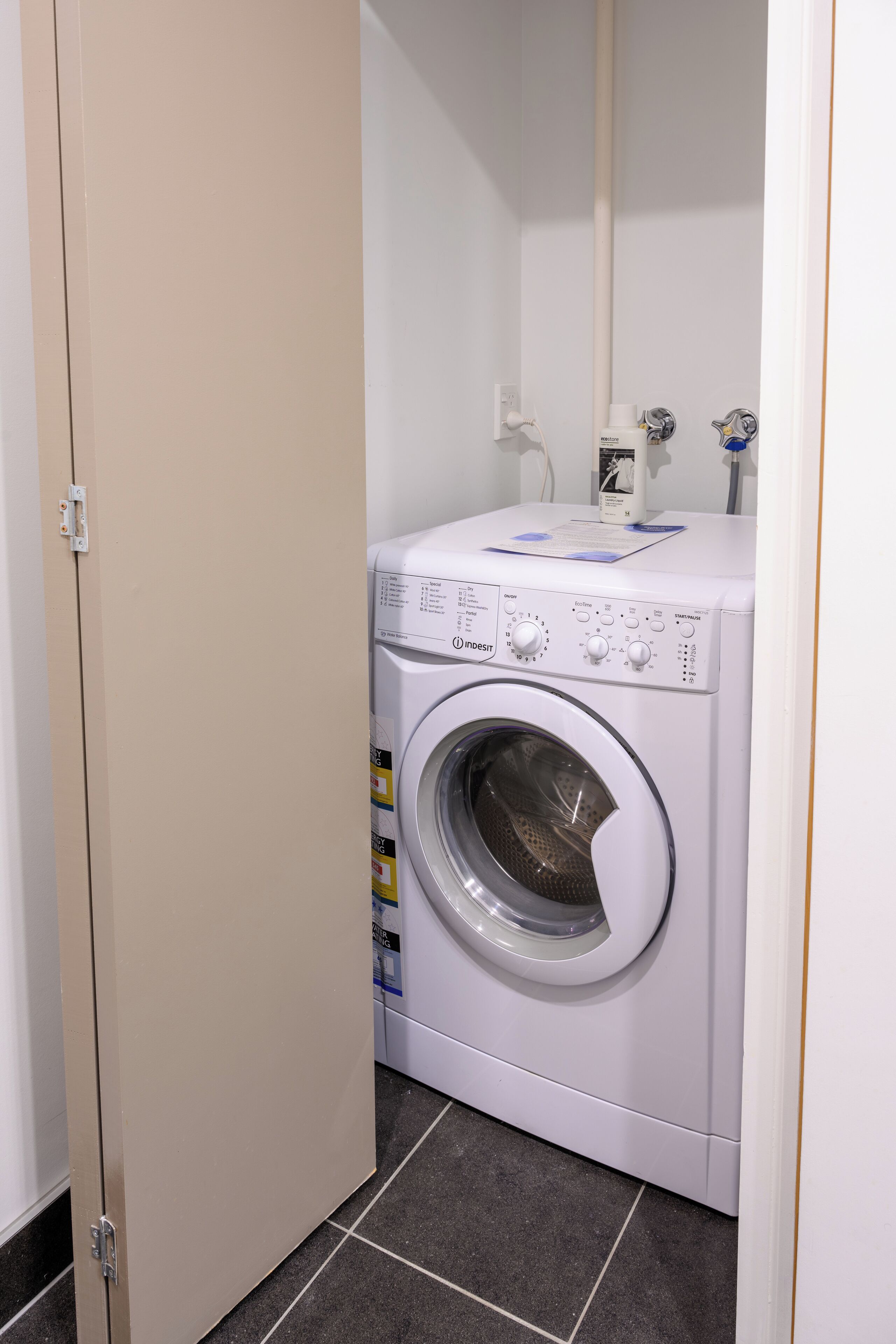 A compact, white front-loading washing machine in a small laundry room with a tiled floor.