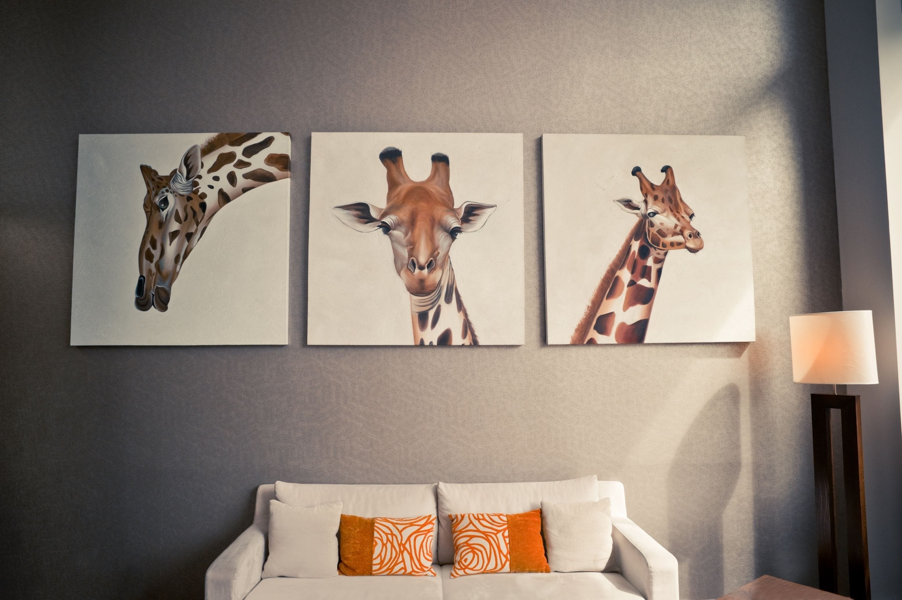 A room with a grey wall, a white sofa with orange patterned pillows, and three giraffe paintings.