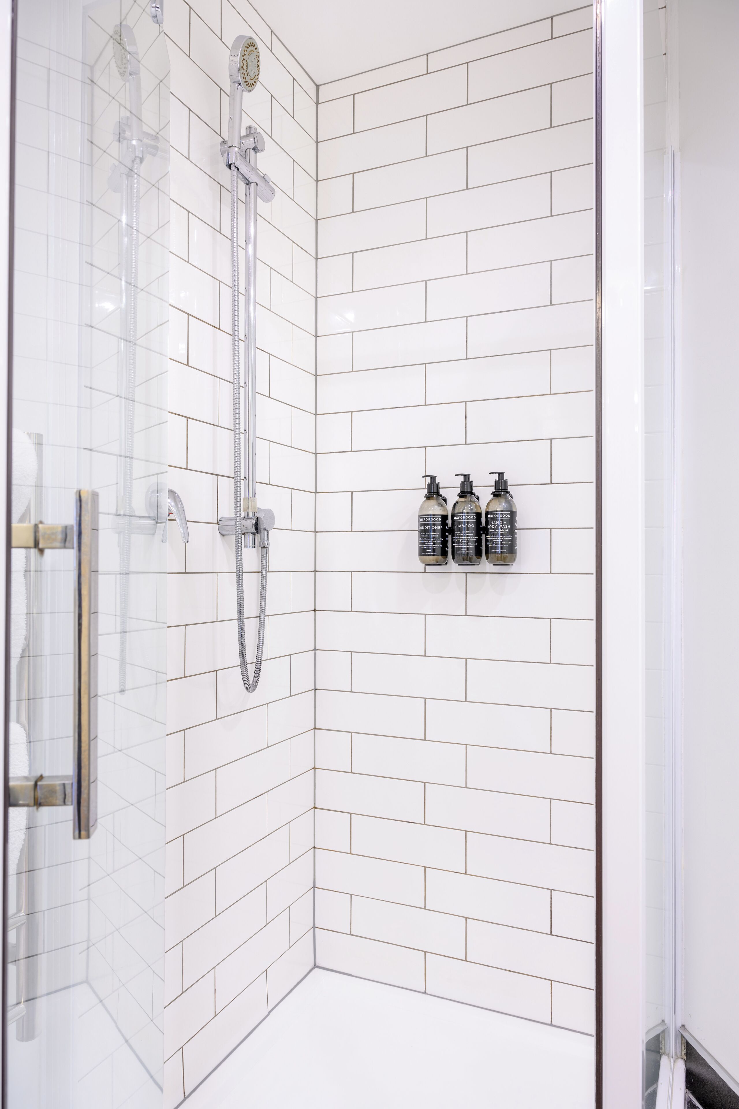 Ducha con puerta de vidrio, cabezal de ducha manual y grifo empotrado en la pared. Hay tres botellas de champú en la pared.
