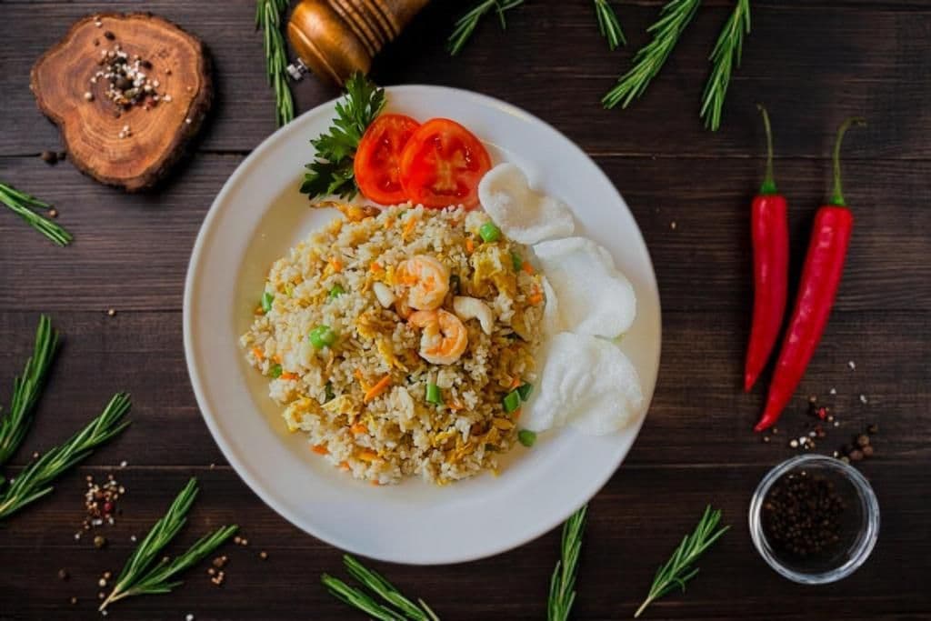 A plate of fried rice with shrimp and vegetables, garnished with tomato slices and herbs, on a dark wooden table with scattered herbs and spices.