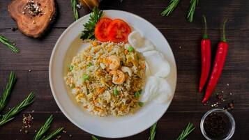 A plate of fried rice with shrimp and vegetables, garnished with tomato slices and herbs, on a dark wooden table with scattered herbs and spices.