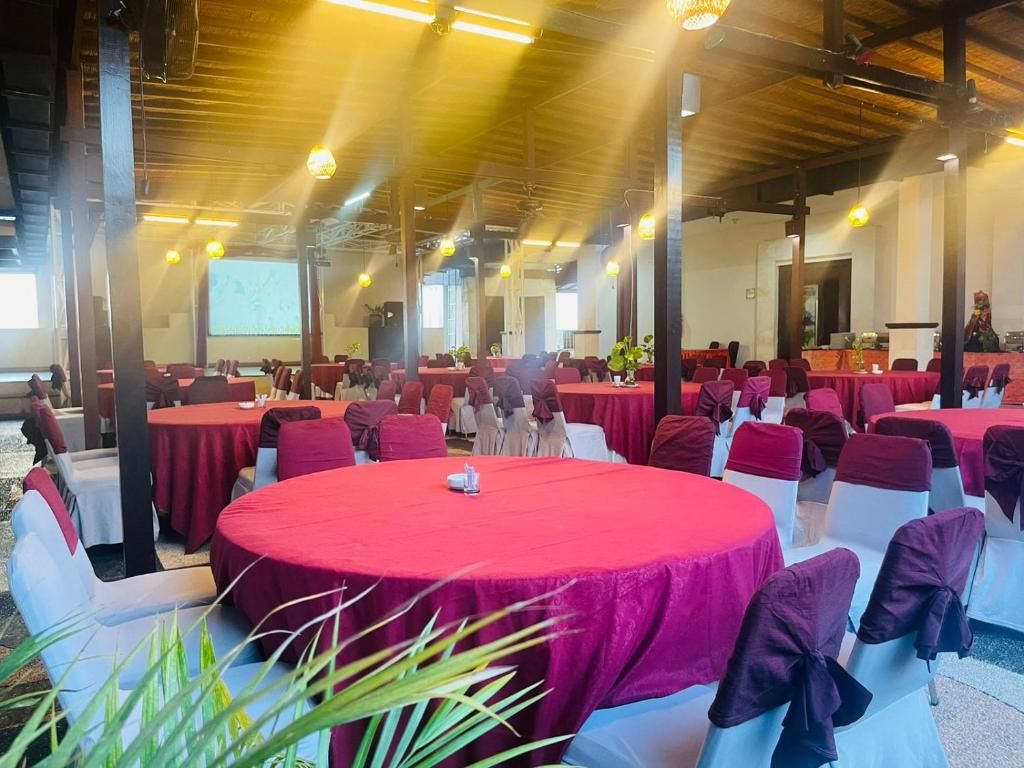 Uma sala de banquetes com mesas redondas cobertas por toalhas vermelhas, cadeiras brancas e um palco.