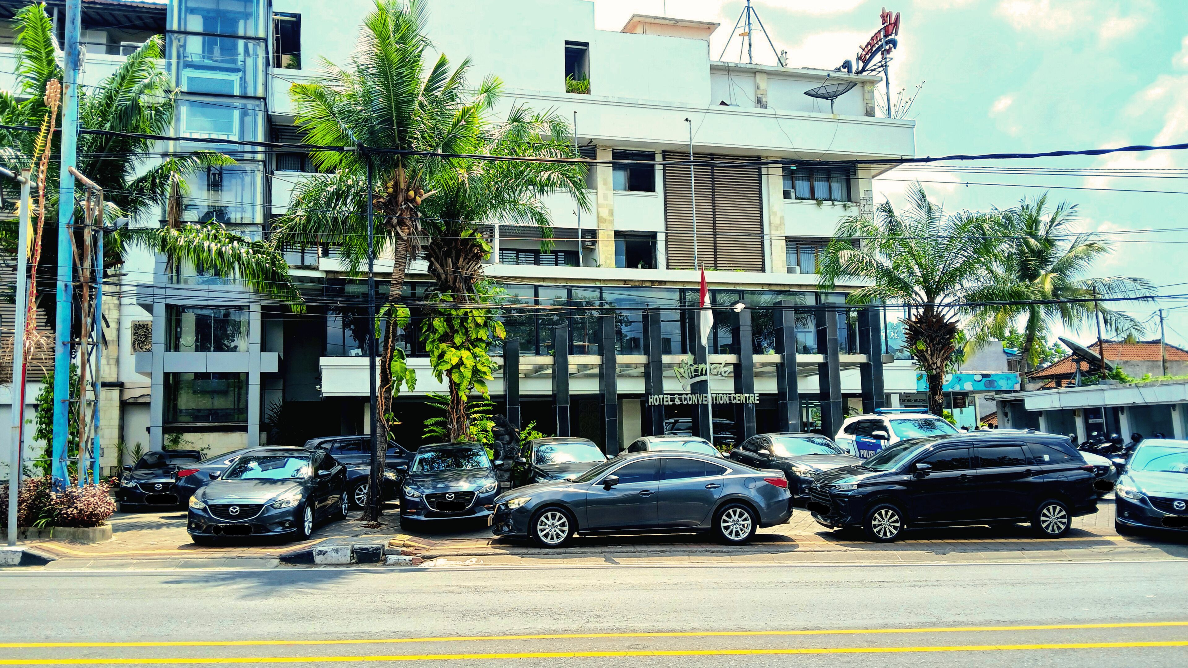 A hotel with a modern glass facade, multiple parking spaces, and palm trees in front.