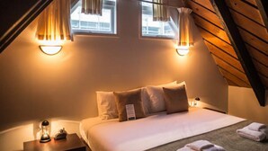A cozy bedroom with a sloped ceiling, two bedside lamps, a bed with white linens, and a small nightstand with a lamp and a clock. - Willis Village (Wellington)