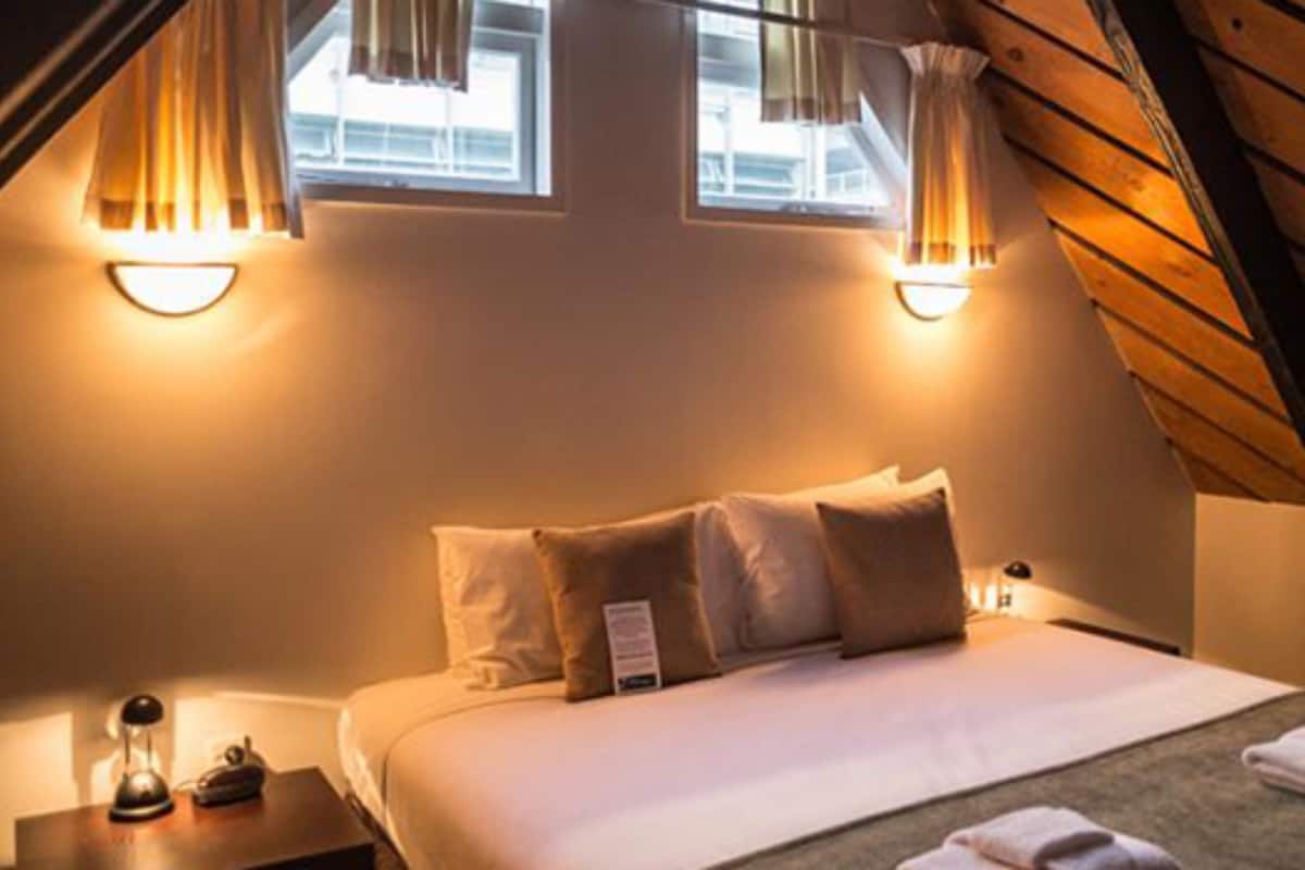 A cozy bedroom with a sloped ceiling, two bedside lamps, a bed with white linens, and a small nightstand with a lamp and a clock.