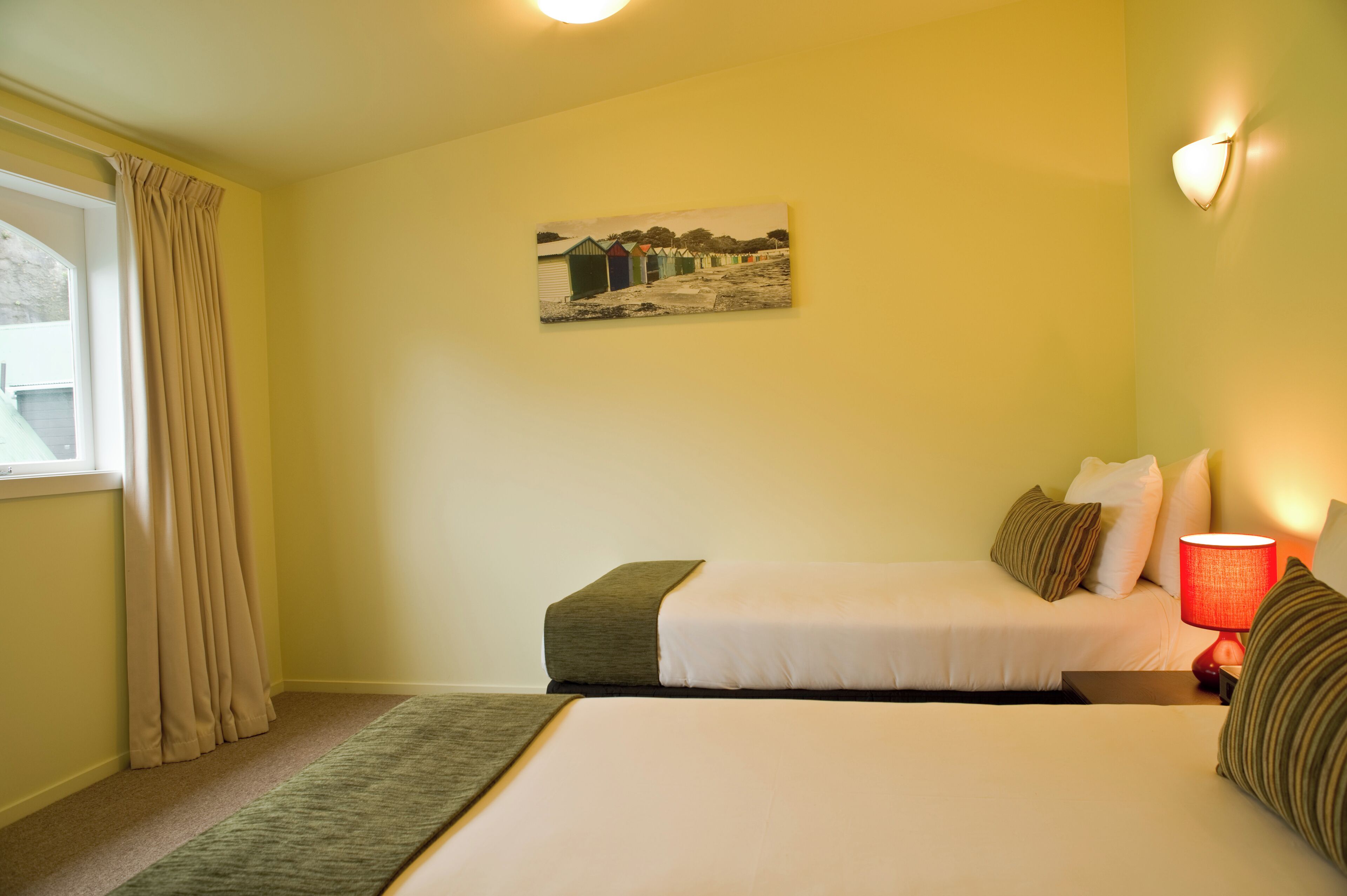 Ein Hotelzimmer mit einem Bett, einem Nachttisch mit Lampe, einem Bild an der Wand und einem Fenster mit Vorhängen.