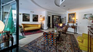 A spacious living room with a yellow sofa, a colorful armchair, a glass coffee table, and a bookshelf. - Hotel Bernardino (Lucca)
