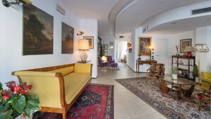 A spacious living room with a yellow sofa, a coffee table, and a patterned rug. There are several chairs and a bookshelf in the background. - Hotel Bernardino (Lucca)