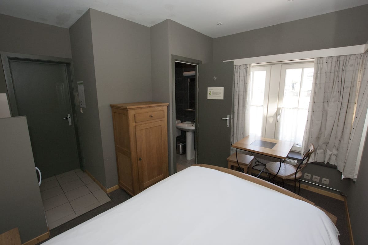 Habitación de hotel con una cama, un armario de madera, un baño con inodoro y lavamanos, y una pequeña mesa con una silla.