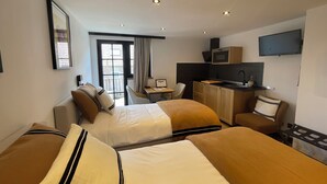 Quarto de hotel com duas camas, uma mesa, cozinha compacta e televisão.
