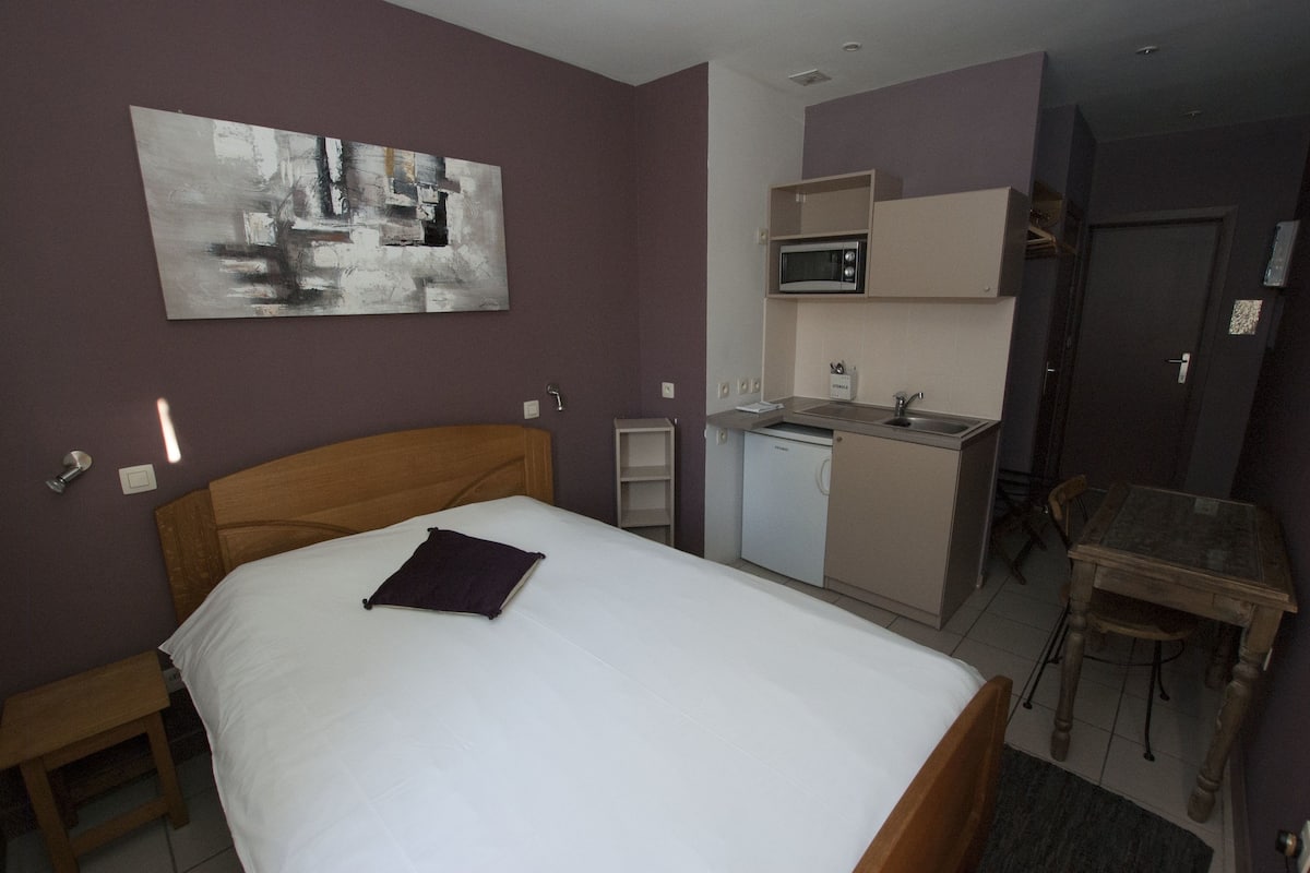 Habitación de hotel con cama, kitchenette pequeña, mesa de comedor y un cuadro en la pared.