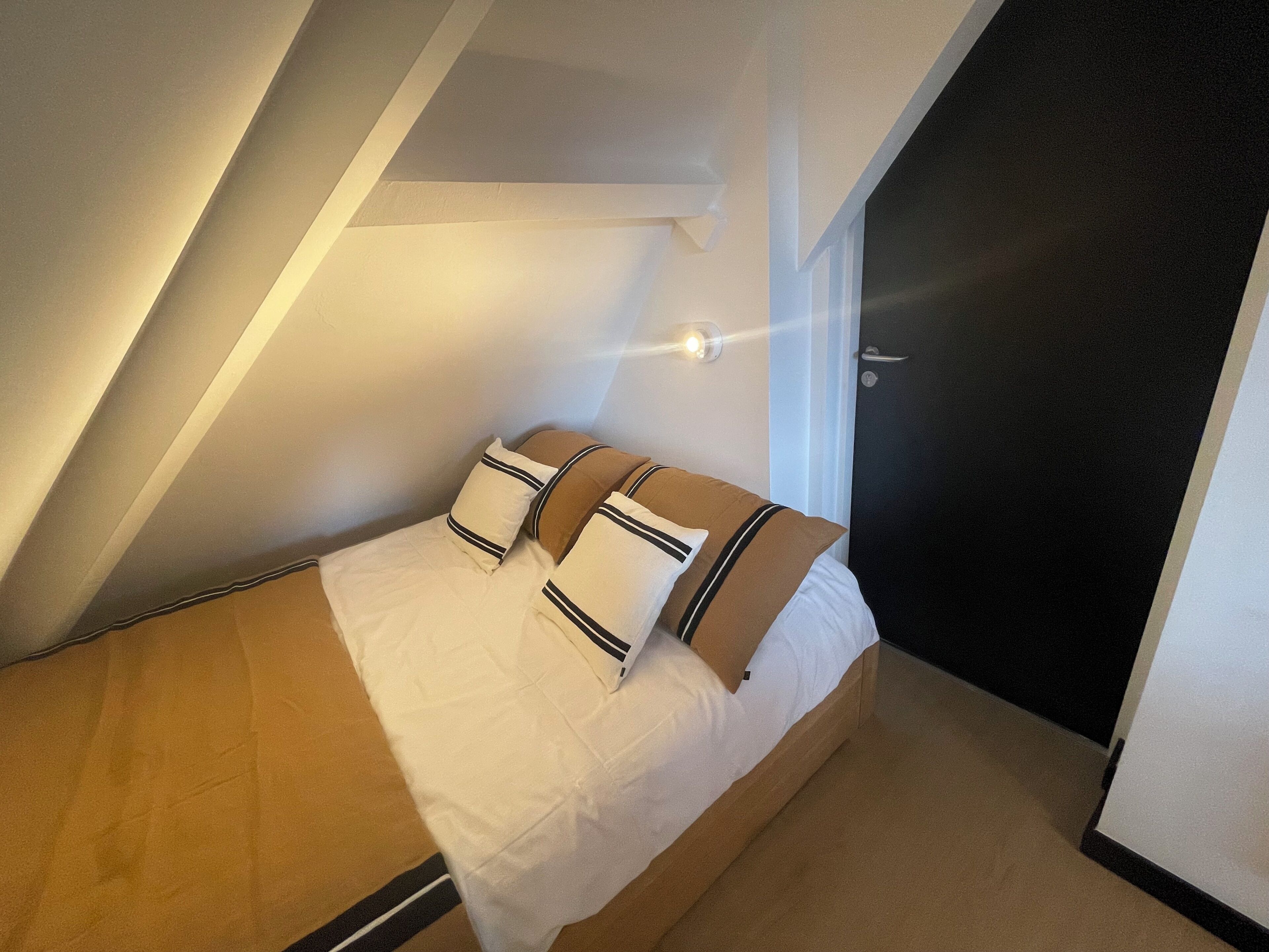 A small, single-bed room with a sloped ceiling, a door, and a wall-mounted light fixture.