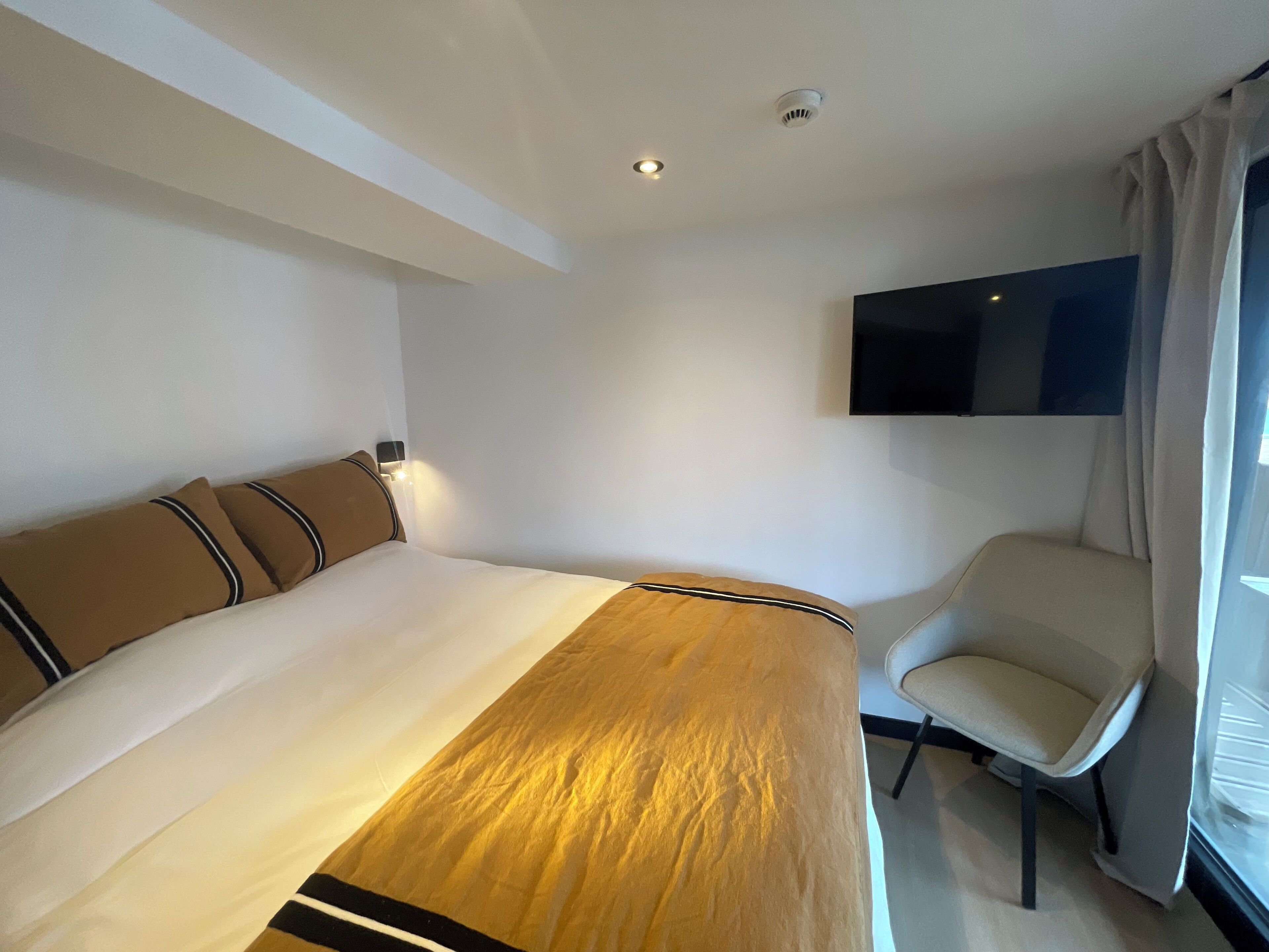 Une chambre d’hôtel avec un lit, une chaise, un téléviseur fixé au mur et une fenêtre avec des rideaux.