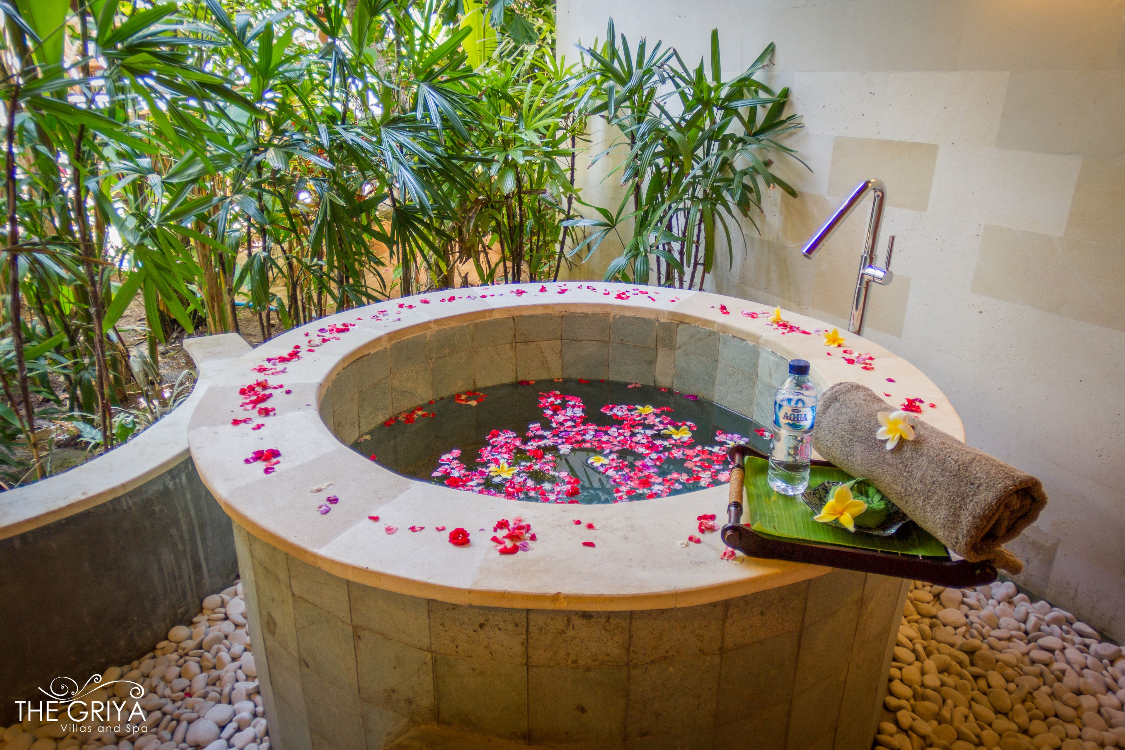 A spa tub surrounded by pebbles and plants, with a towel and flowers on a tray.