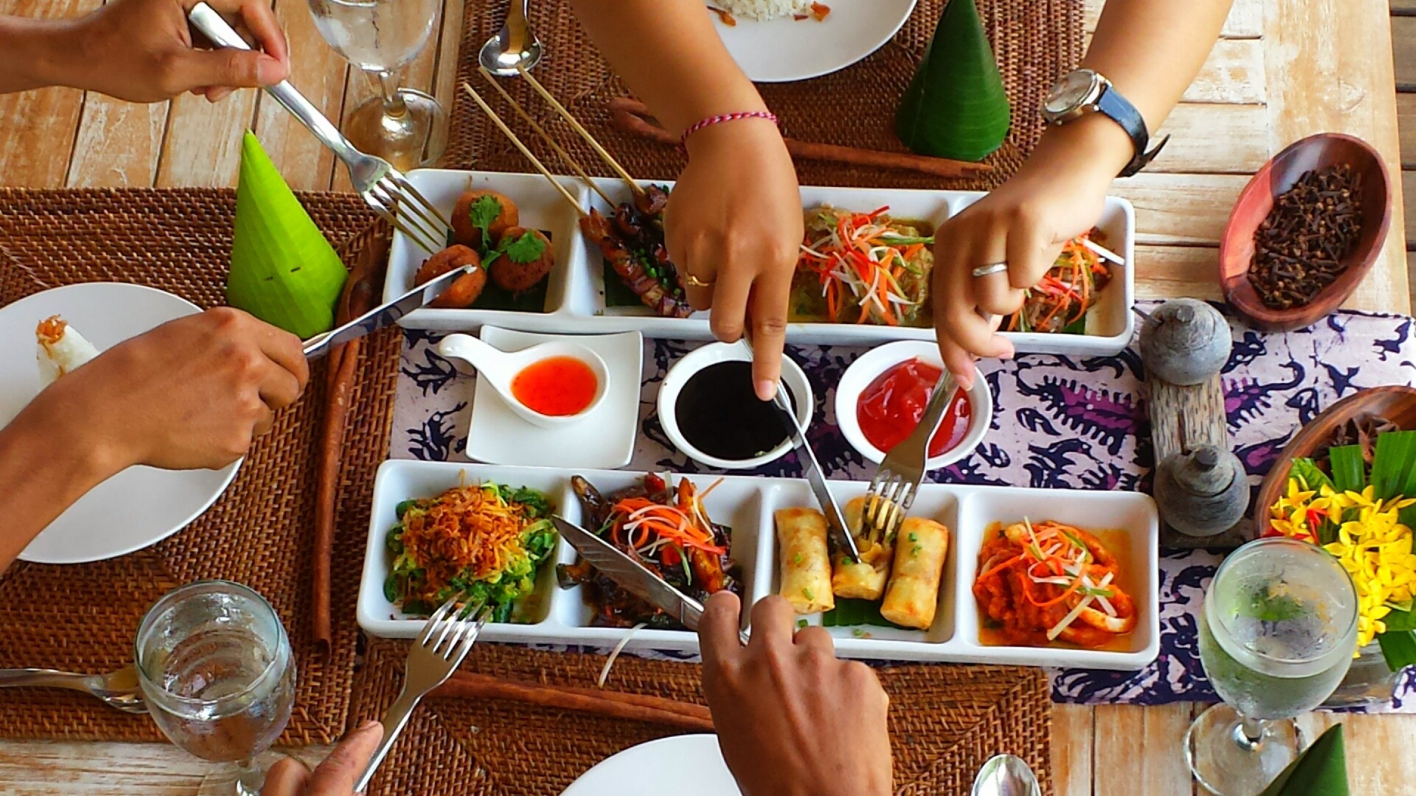 A dining table with various dishes, including spring rolls, salads, and sauces, with people using forks and chopsticks.