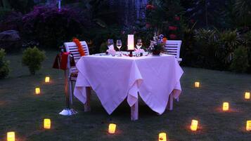 Un área de comedor al aire libre con una mesa preparada para dos personas, rodeada de velas y plantas tropicales.