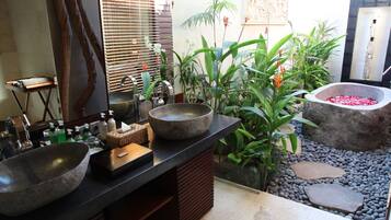 A bathroom with a large stone sink, a wooden vanity, and a stone basin filled with water and rose petals. There are various plants and a mirror on the wall.