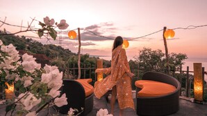 A woman in a floral dress standing on a balcony overlooking a sunset, with wicker furniture and string lights. - The Griya Villas and Spa (Karangasem)