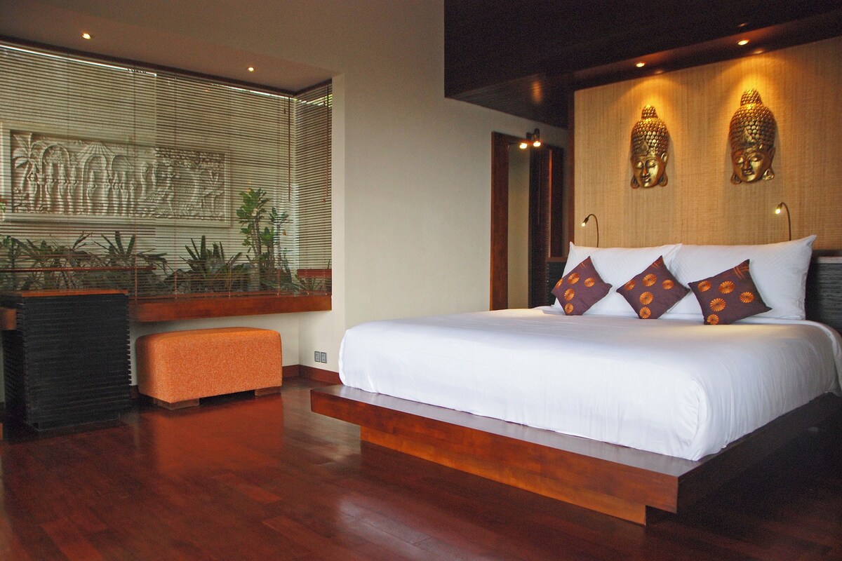 A modern bedroom with a large bed, wooden flooring, a bench, and a wall with Buddha statues.
