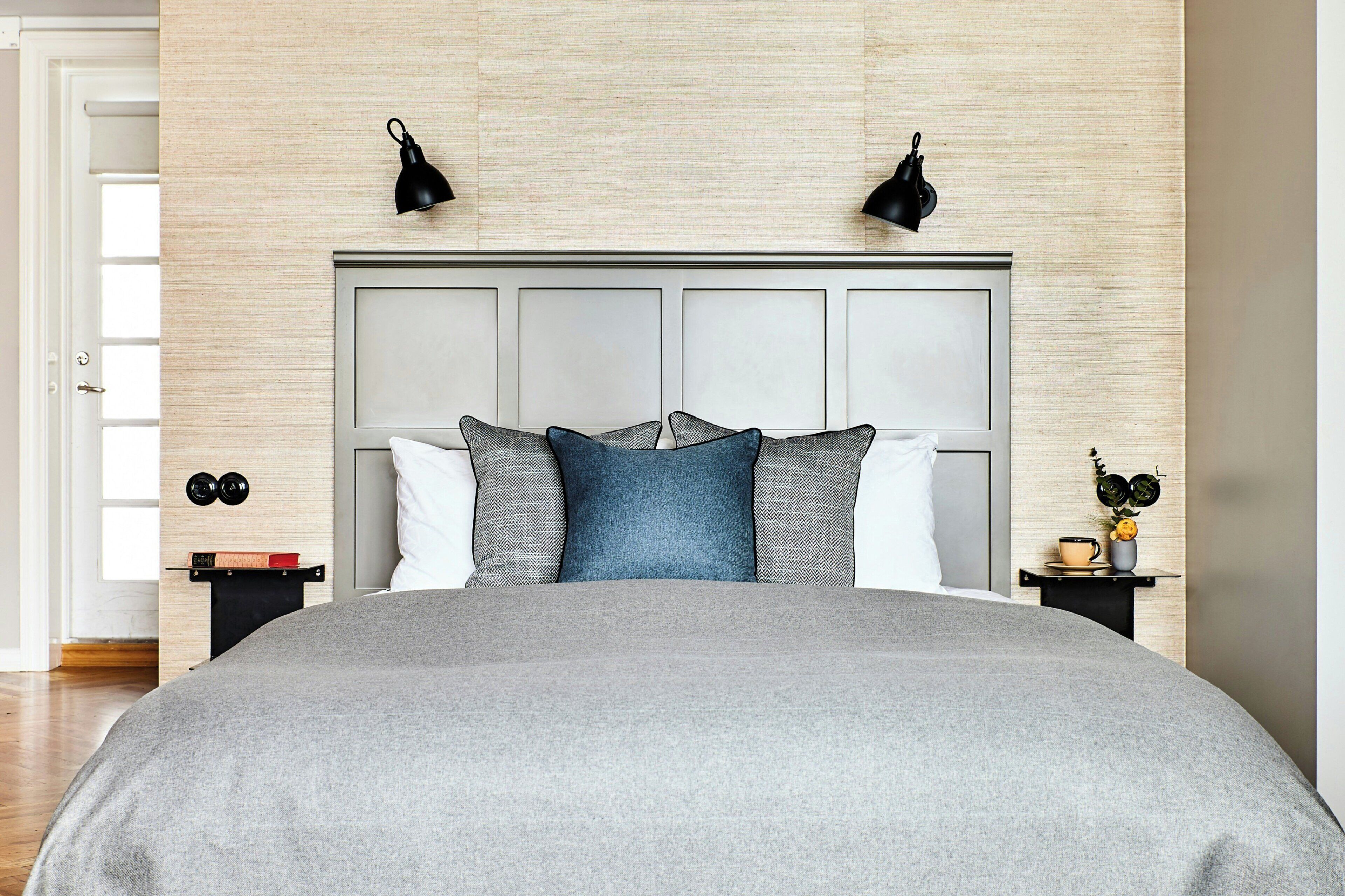 A bedroom with a large bed, a headboard with panels, two bedside lamps, a nightstand with a book and a clock, and a small table with a cup and a flower.