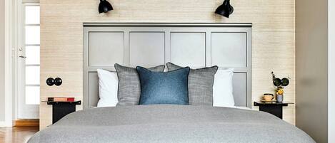 A bedroom with a large bed, a headboard with panels, two bedside lamps, a nightstand with a book and a clock, and a small table with a cup and a flower.