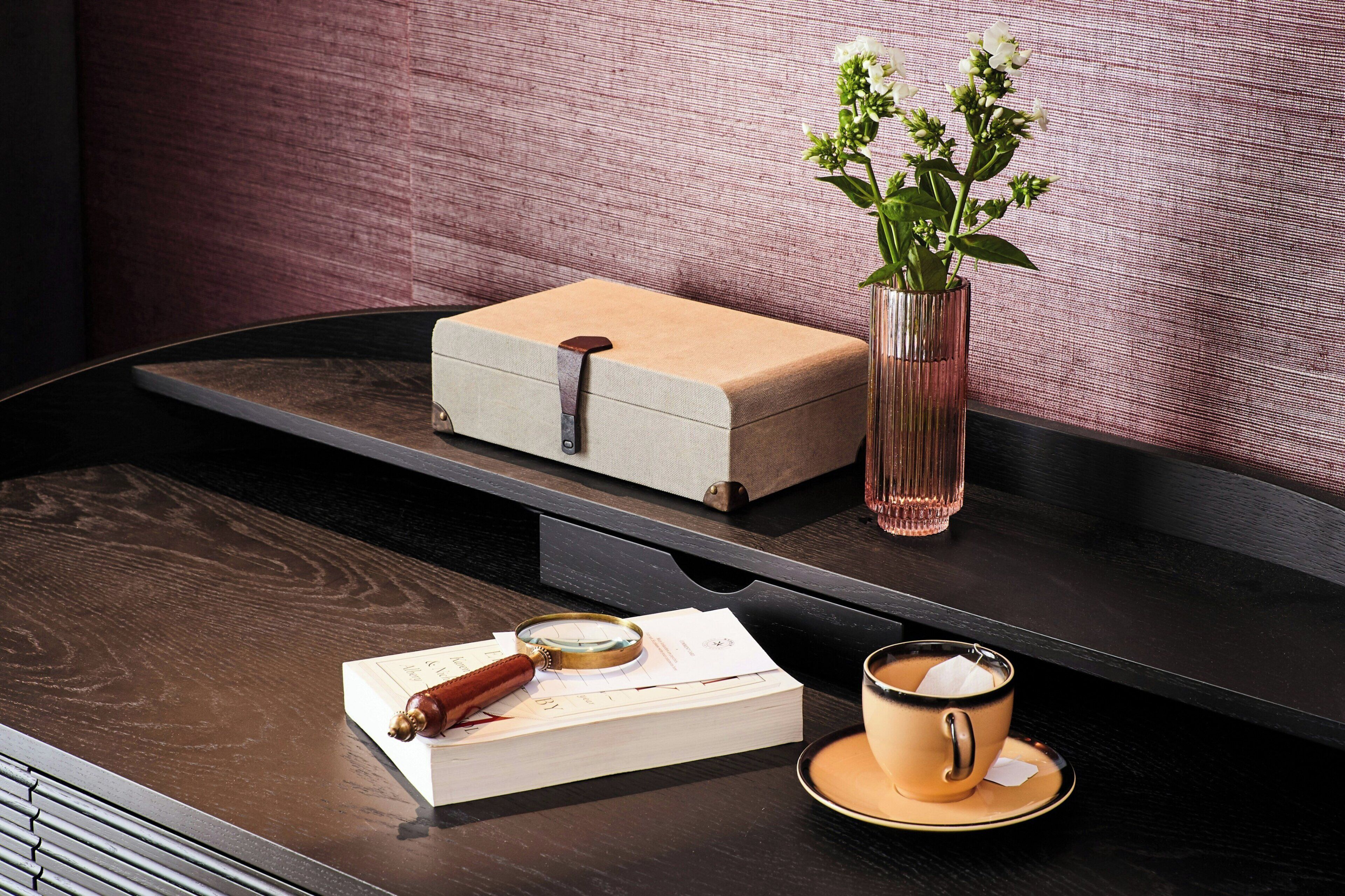 A wooden table with a white box, a magnifying glass, a book, a vase with flowers, and a cup of coffee.
