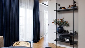 A modern living room with a dining area, featuring a dark blue curtain, a black standing lamp, a shelf with books and a vase of flowers, and a wooden floor. - Kvosin Downtown Hotel (Reykjavik)