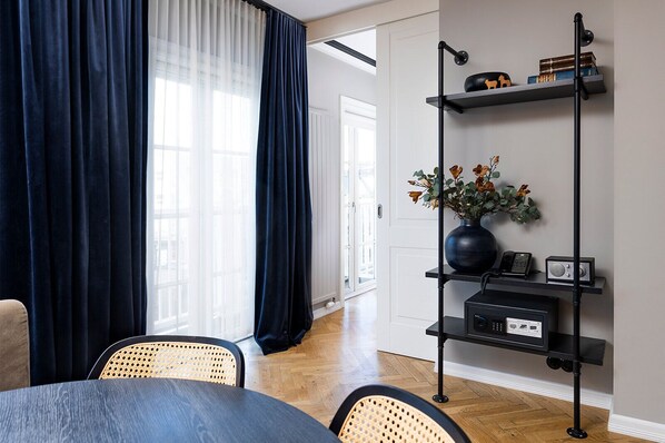Ein modernes Wohnzimmer mit Essbereich, mit einem dunkelblauen Vorhang, einer schwarzen Stehlampe, einem Regal mit Büchern und einer Vase mit Blumen sowie einem Holzboden.
