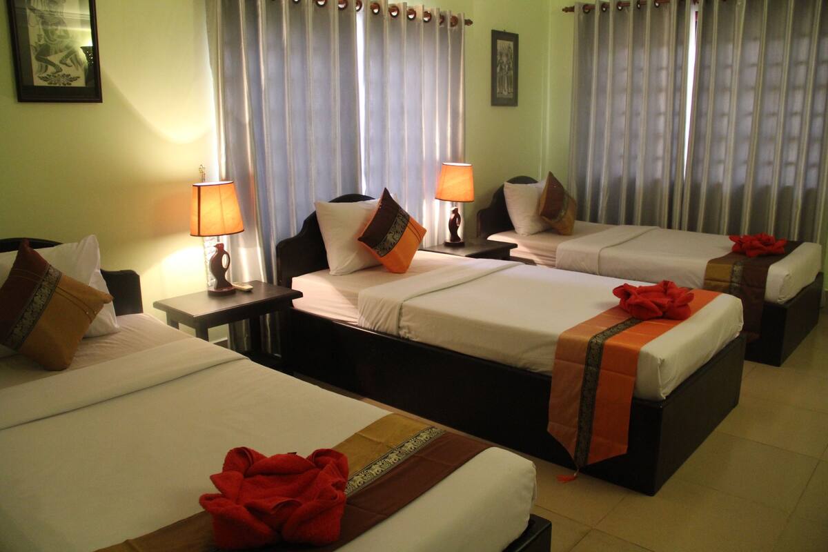 Habitación de hotel con tres camas individuales, cada una con almohada y manta. Hay dos mesitas de noche con lámparas. La habitación tiene cortinas y un cuadro en la pared.