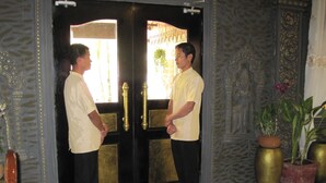 Two men standing in front of a black door with brass handles, a checkered floor, and a potted plant to the right. - Popular Boutique Hotel (Siem Reap)