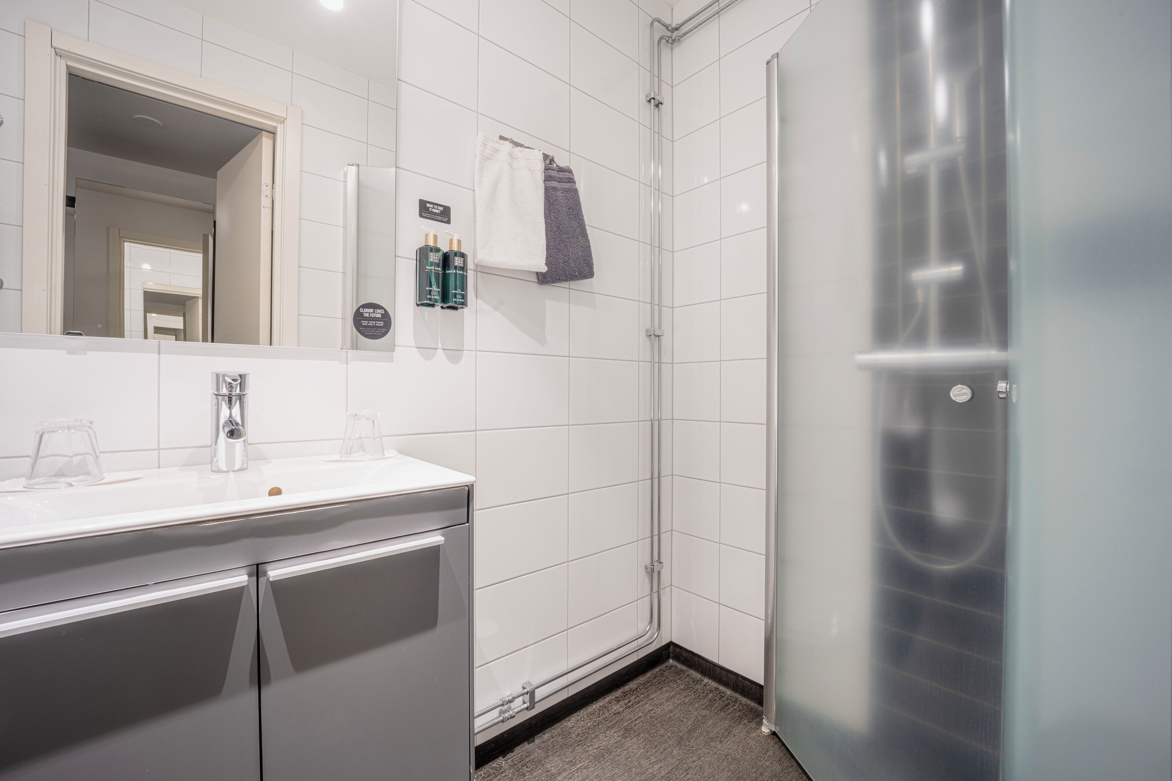 Une salle de bain moderne avec un mur recouvert de carreaux blancs, une douche en verre, un lavabo blanc avec un miroir et un porte-serviettes avec une serviette.