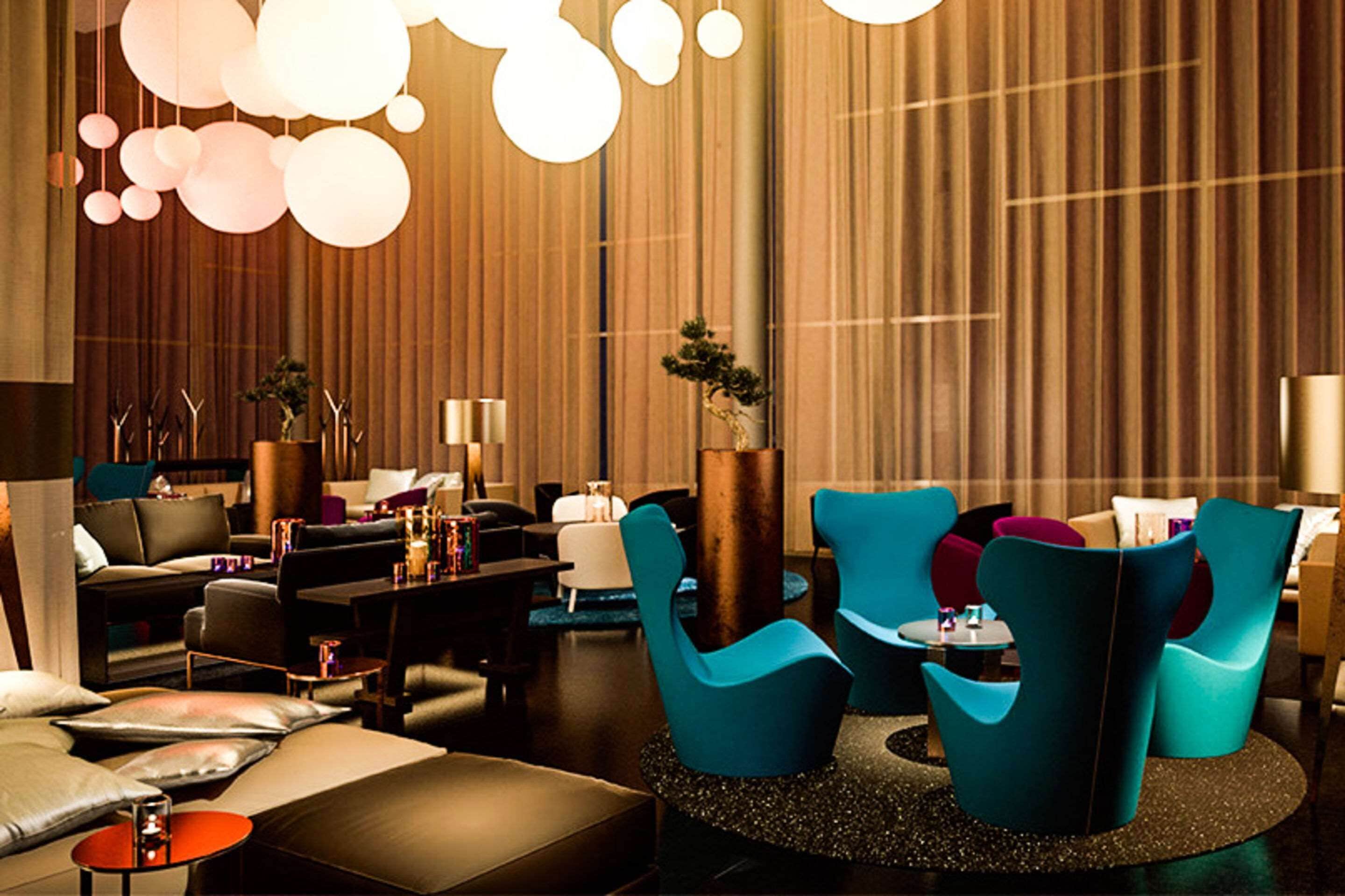 A modern hotel lobby with a seating area featuring teal chairs, a sofa, and a coffee table. There are decorative plants and a large, round table with a lamp.