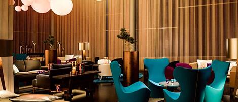 A modern hotel lobby with a seating area featuring teal chairs, a sofa, and a coffee table. There are decorative plants and a large, round table with a lamp.