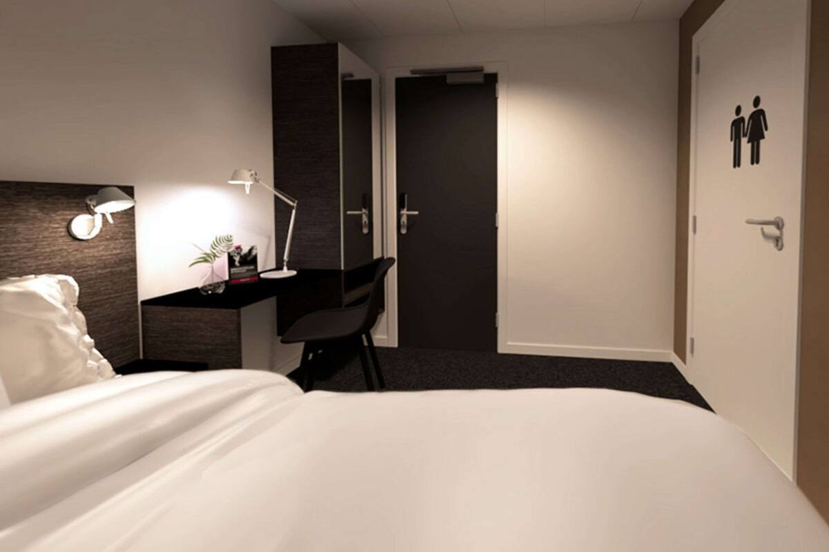 A hotel room with a bed, a desk with a chair, a lamp, a small plant, and a door with a male and female restroom sign.