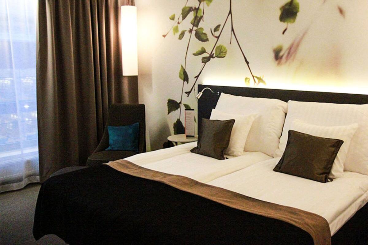 A hotel room with a large bed, a chair, a bedside table, and a view of the city through the window.