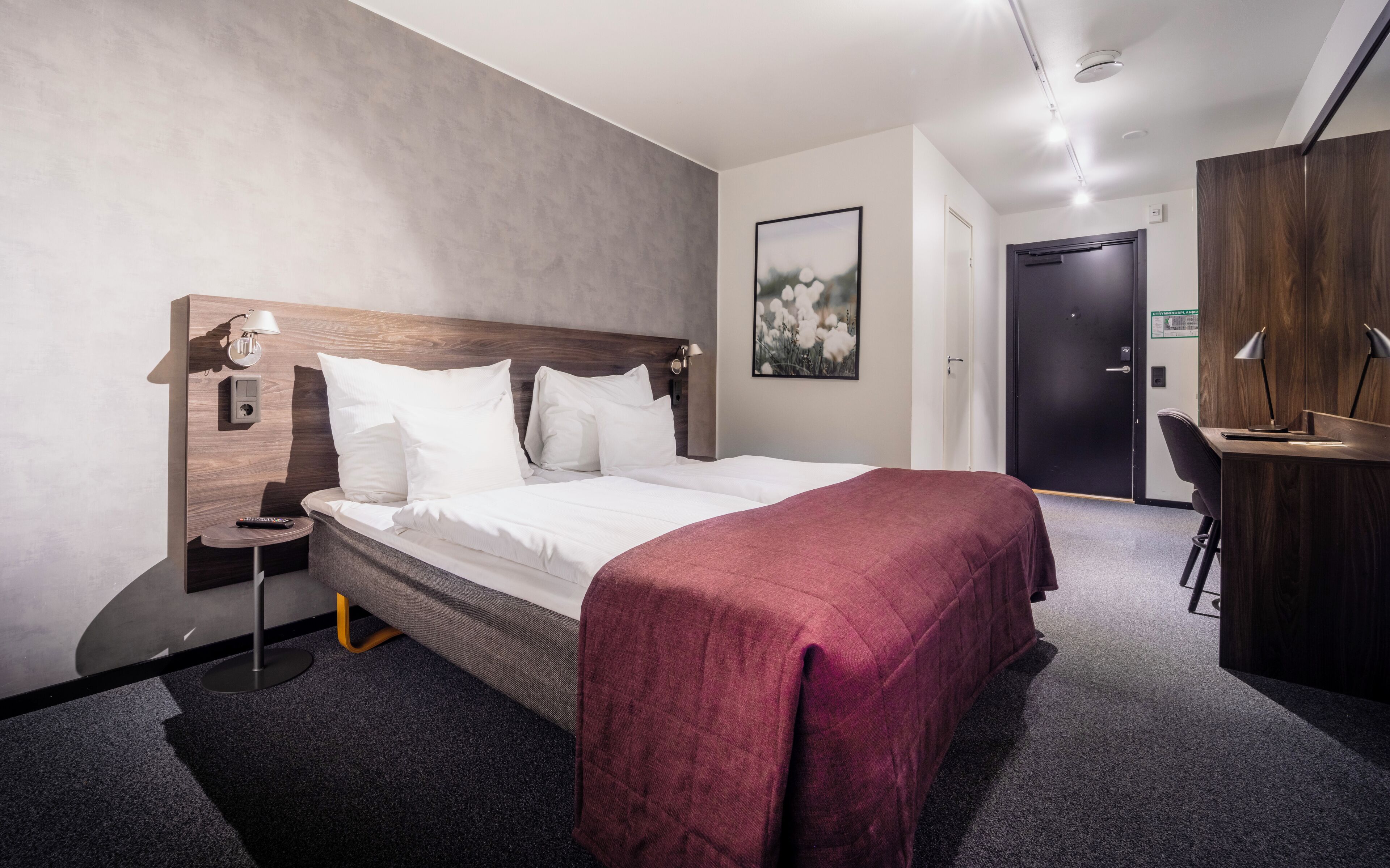 A hotel room with a bed, bedside table, lamp, and a desk with a chair.