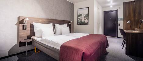 A hotel room with a bed, bedside table, lamp, and a desk with a chair.