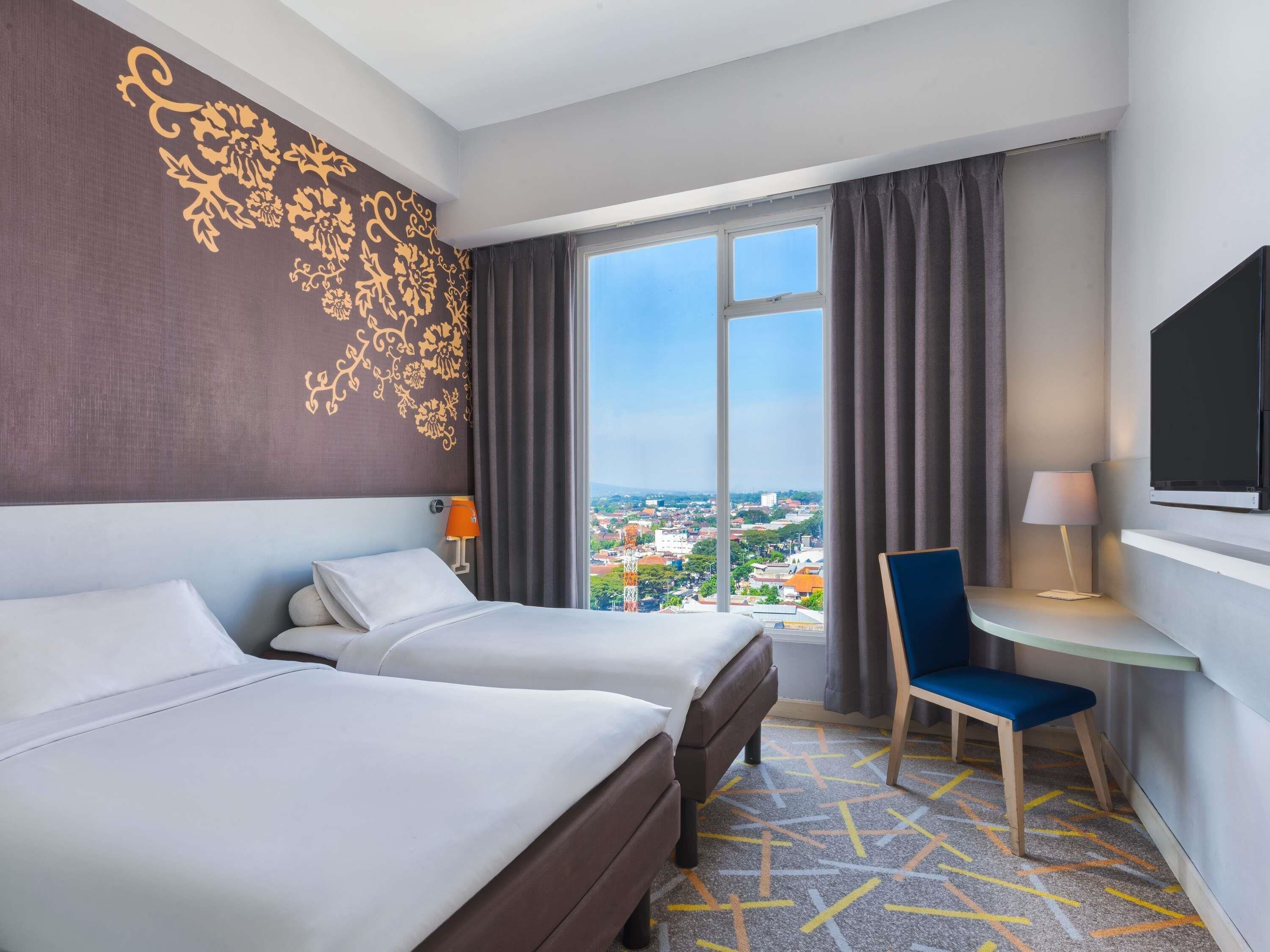 A hotel room with two beds, a desk, a chair, a TV, and a large window with a city view.