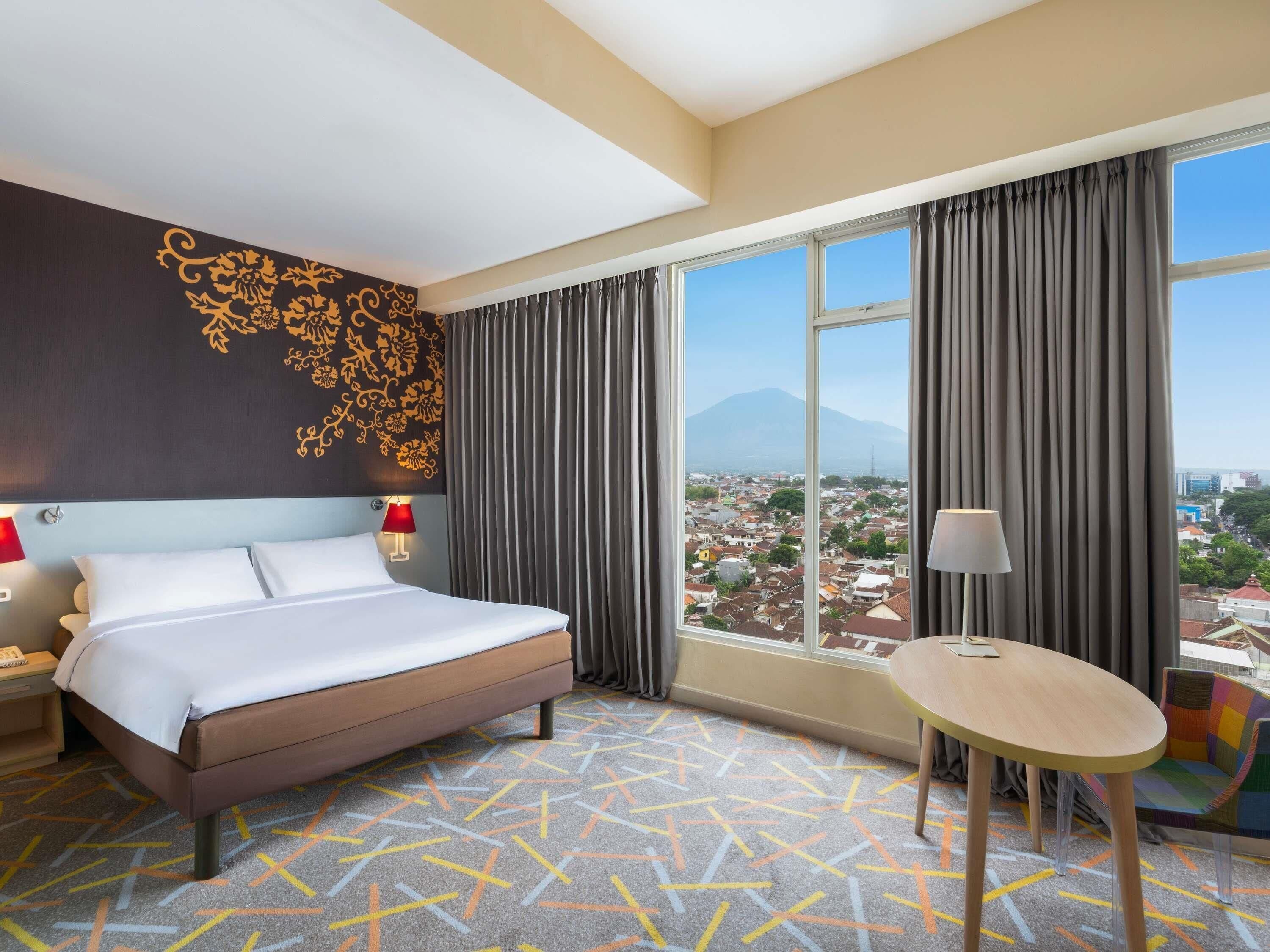 A hotel room with a bed, a desk, a chair, a view of a cityscape, and a decorative wall.
