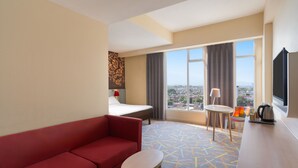 A hotel room with a red sofa, a small round table, a bed, a TV, and a view of the city. - Ibis Styles Malang (Malang)
